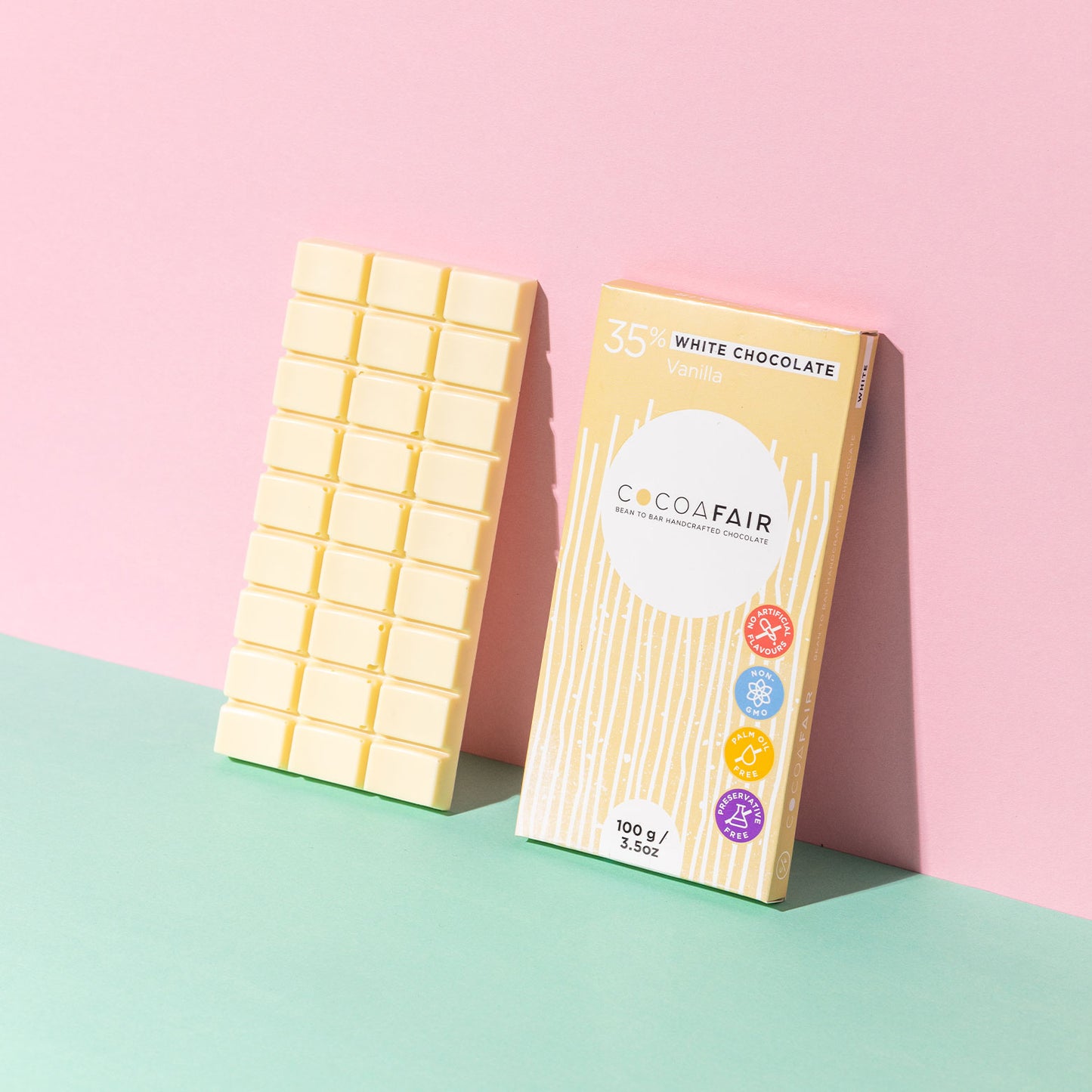 COCOAFAIR 100g 35% White Chocolate Lifestyle shot - Cream packaging with a white chococolate slab on pink and green background at a angle. Vanilla flavoured chocolate.