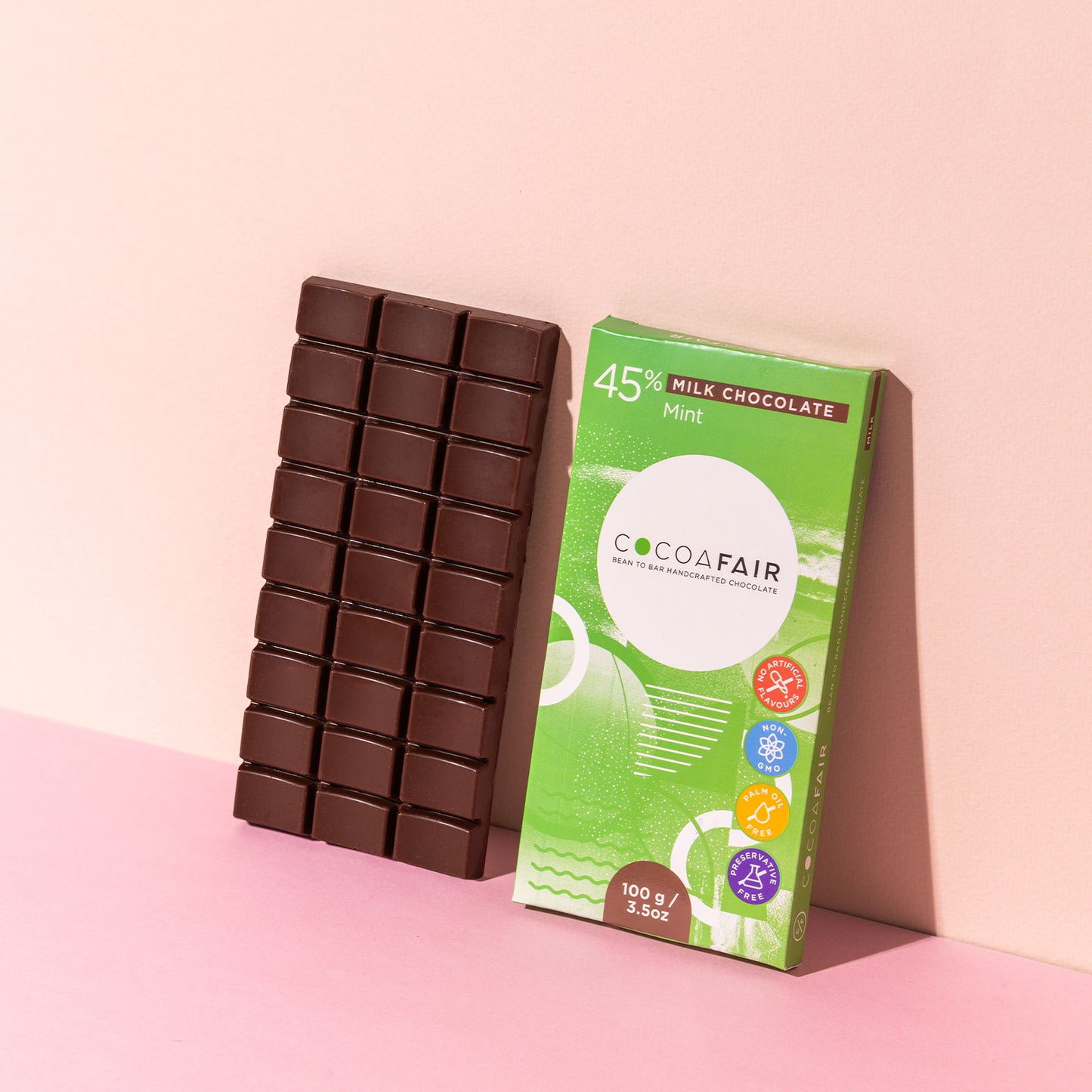 COCOAFAIR 100g 45% Milk Chocolate Lifestyle shot - Green packaging with milk chocolate slab on pink background at a angle. Mint flavoured chocolate.